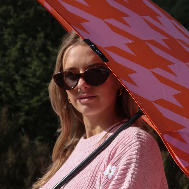Woman holding a red umbrella with a pattern, wearing sunglasses and a pink sweater.