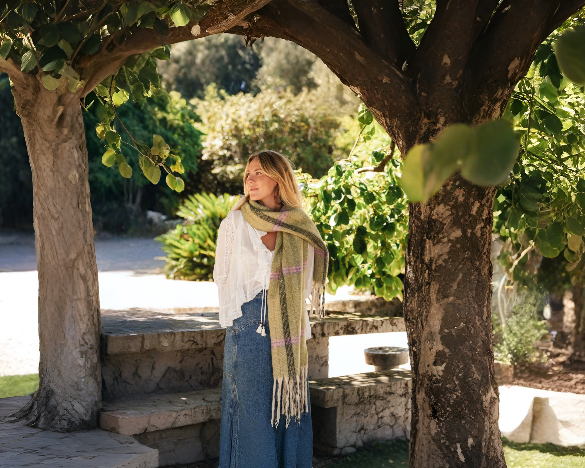 A woman standing outside among trees wearing a green scarf, white top and blue jeans