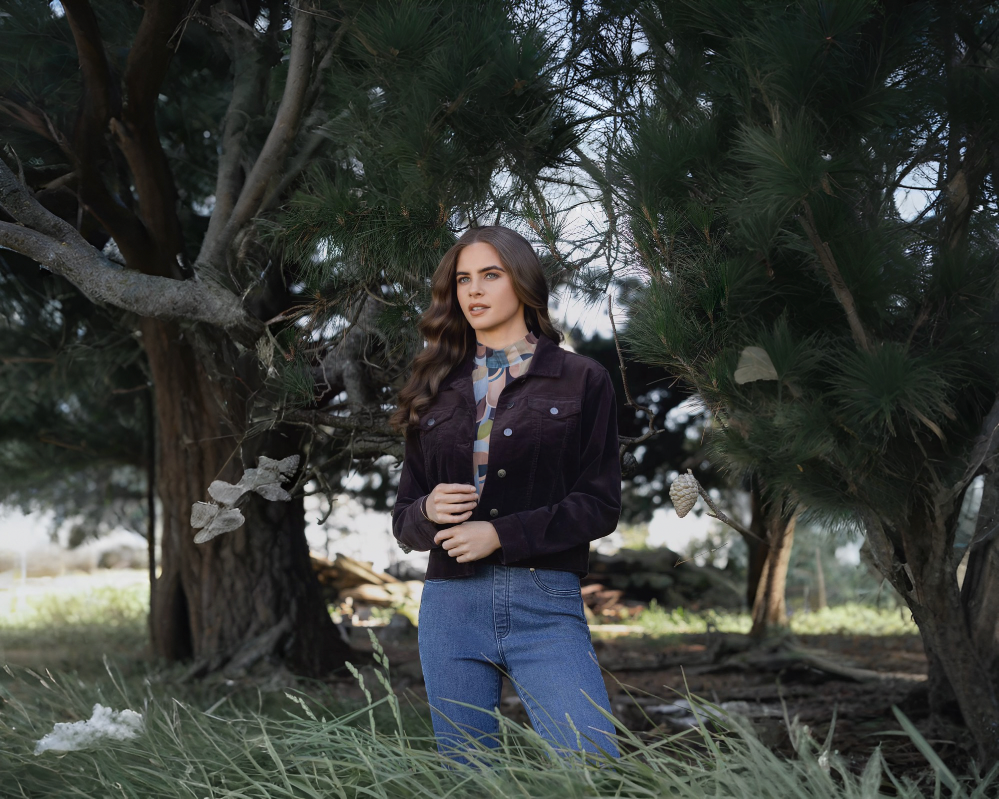 A woman standing in long grass wearing a dark purple jacket and blue jeans